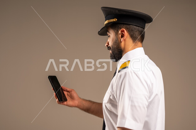 استخدام الاجهزة التقنية الحديثة، طاقم الاستقبال والترحيب بالركاب، العمل في مجال الطيران، مفهوم السياحة والسفر، رحلات جوية سياحية, خطوط الطيران السعودية,  بورتريه لكابتن طيار عربي سعودي خليجي مبتسم يرتدي الزي الرسمي يمسك بيده الهاتف المحمول، خلفية ملونة
