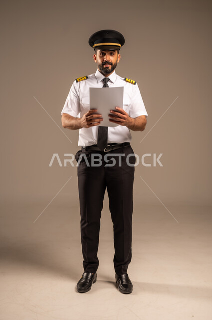 القاء ارشادات السلامة على المسافرين، العمل في مجال الطيران، طاقم الاستقبال والترحيب بالركاب، مفهوم السياحة والسفر، رحلات جوية سياحية, خطوط الطيران السعودية,  بورتريه لكابتن طيار عربي سعودي خليجي مبتسم يرتدي الزي الرسمي يحمل بيده ورقة بيضاء فارغة، خلفية ملونة