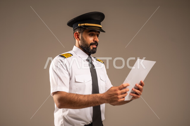 القاء ارشادات السلامة على المسافرين، العمل في مجال الطيران، طاقم الاستقبال والترحيب بالركاب، مفهوم السياحة والسفر، رحلات جوية سياحية, خطوط الطيران السعودية,  بورتريه لكابتن طيار عربي سعودي خليجي مبتسم يرتدي الزي الرسمي يحمل بيده ورقة بيضاء فارغة، خلفية ملونة
