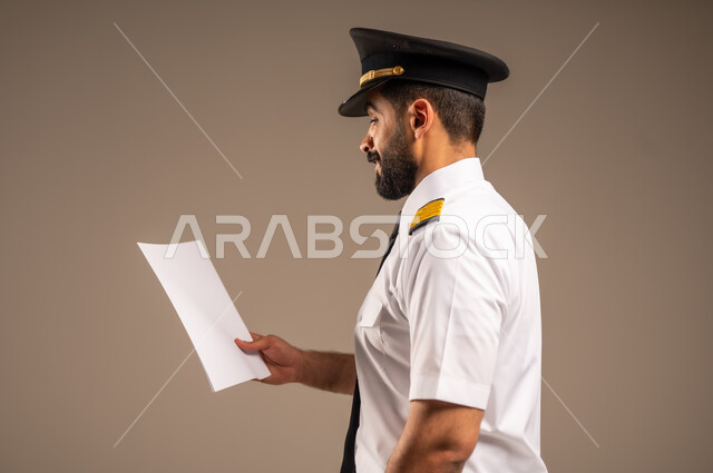 القاء ارشادات السلامة على المسافرين، العمل في مجال الطيران، طاقم الاستقبال والترحيب بالركاب، مفهوم السياحة والسفر، رحلات جوية سياحية, خطوط الطيران السعودية,  بورتريه لكابتن طيار عربي سعودي خليجي مبتسم يرتدي الزي الرسمي يحمل بيده ورقة بيضاء فارغة، خلفية ملونة