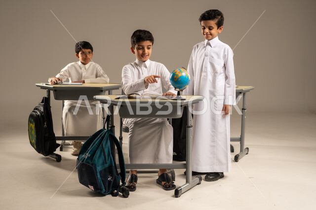 The concept of exploration and research around the world, the development of teaching methods in the Kingdom’s schools, the quality of effective learning methods and the use of tools and aids, a portrait of Saudi Arabian Gulf students wearing school uniforms sitting in their seats with a miniature globe in front of them, gray background