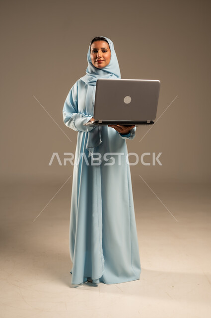 ادارة الأعمال عن بعد، استخدام الأجهزة الحديثة والمتطورة، بورتريه لامرأة عربية خليجية سعودية ترتدي العباءة والحجاب تقوم باستخدام الحاسوب المحمول، دمج التكنولوجيا بالعمل، إنجاز المهام المطلوبة عبر اللابتوب، مهن ووظائف نسائية، خلفية ملونة