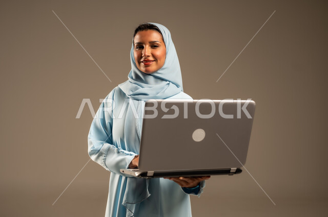 Remote business management, use of modern and advanced devices, portrait of a Saudi Arabian Gulf woman wearing an abaya and hijab using a laptop, integrating technology into work, completing required tasks via laptop, women's professions and jobs, colorful background
