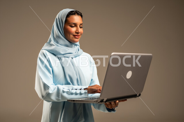 ادارة الأعمال عن بعد، استخدام الأجهزة الحديثة والمتطورة، بورتريه لامرأة عربية خليجية سعودية ترتدي العباءة والحجاب تقوم باستخدام الحاسوب المحمول، دمج التكنولوجيا بالعمل، إنجاز المهام المطلوبة عبر اللابتوب، مهن ووظائف نسائية، خلفية ملونة