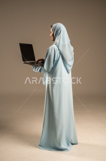 ادارة الأعمال عن بعد، استخدام الأجهزة الحديثة والمتطورة، بورتريه لامرأة عربية خليجية سعودية ترتدي العباءة والحجاب تقوم باستخدام الحاسوب المحمول، دمج التكنولوجيا بالعمل، إنجاز المهام المطلوبة عبر اللابتوب، مهن ووظائف نسائية، خلفية ملونة