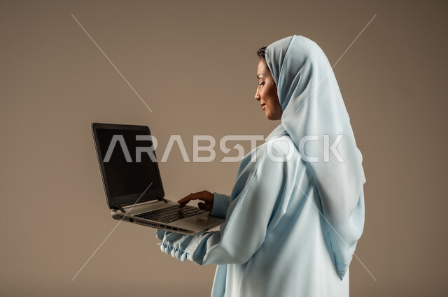 ادارة الأعمال عن بعد، استخدام الأجهزة الحديثة والمتطورة، بورتريه لامرأة عربية خليجية سعودية ترتدي العباءة والحجاب تقوم باستخدام الحاسوب المحمول، دمج التكنولوجيا بالعمل، إنجاز المهام المطلوبة عبر اللابتوب، مهن ووظائف نسائية، خلفية ملونة