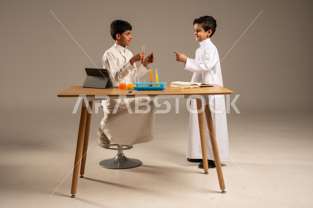 Teamwork and cooperation between colleagues, taking advantage of technology and technological devices in effective learning, using experimental educational tools and means, portrait of two Saudi Arabian Gulf students wearing school uniforms conducting chemical experiments and tests, gray background