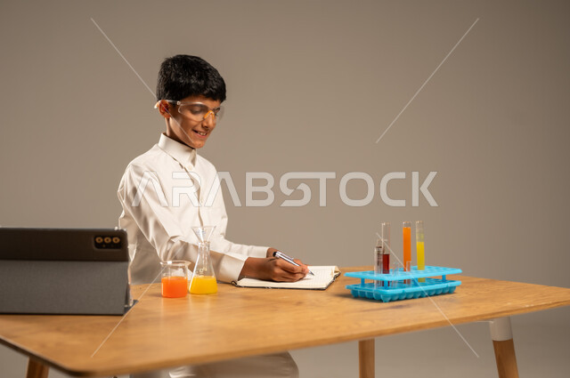 Utilizing modern technological techniques and devices for effective learning, using experimental educational tools and means, portrait of a Saudi Arabian Gulf student wearing school uniform and safety glasses sitting behind an examination table and sample tubes and chemical analyses, gray background