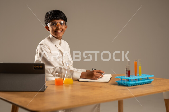 Utilizing modern technological techniques and devices for effective learning, using experimental educational tools and means, portrait of a Saudi Arabian Gulf student wearing school uniform and safety glasses sitting behind an examination table and sample tubes and chemical analyses, gray background