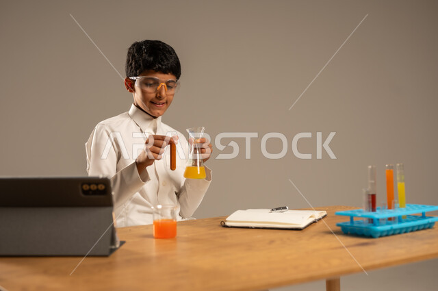 Utilizing modern technological techniques and devices for effective learning, using experimental educational tools and means, portrait of a Saudi Arabian Gulf student wearing school uniform and safety glasses sitting behind an examination table and sample tubes and chemical analyses, gray background