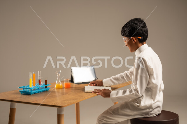 Using experimental teaching tools and methods, portrait from the back of a Saudi Arabian Gulf student wearing school uniform and safety glasses sitting behind a test table and sample tubes and chemical analyses, taking advantage of technology and technological devices in effective learning, gray background