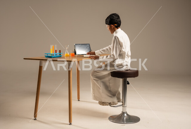 استخدام الأدوات والوسائل التعليمية التجريبية، بورتريه من الخلف لطالب عربي خليجي سعودي يرتدي الزي المدرسي ونظارات الحماية يجلس خلف طاولة الاختبارات وأنابيب العينات والتحاليل الكيميائية، الاستفادة من التقنيات والأجهزة التكنولوجية بالتعلم الفعال، خلفية رمادية
