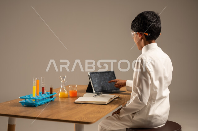 Using experimental teaching tools and methods, portrait from the back of a Saudi Arabian Gulf student wearing school uniform and safety glasses sitting behind a test table and sample tubes and chemical analyses, taking advantage of technology and technological devices in effective learning, gray background