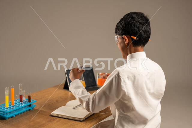 استخدام الأدوات والوسائل التعليمية التجريبية، بورتريه من الخلف لطالب عربي خليجي سعودي يرتدي الزي المدرسي ونظارات الحماية يجلس خلف طاولة الاختبارات وأنابيب العينات والتحاليل الكيميائية، الاستفادة من التقنيات والأجهزة التكنولوجية بالتعلم الفعال، خلفية رمادية