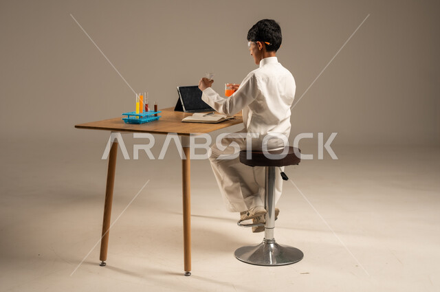 Using experimental teaching tools and methods, portrait from the back of a Saudi Arabian Gulf student wearing school uniform and safety glasses sitting behind a test table and sample tubes and chemical analyses, taking advantage of technology and technological devices in effective learning, gray background