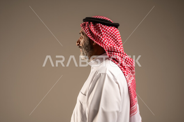 النظر الى شيء ما بإيماءات الجدية، التقدم بالعمر والهيبة والوقار، بورتريه مقرب من الجانب لرجل كبير بالسن عربي خليجي سعودي يرتدي الثوب التقليدي و الشماغ, الاهتمام بالأناقة والمظهر الخارجي، الوقوف باستقامة وثبات، مفهوم الرجولة العربية, خلفية بيج