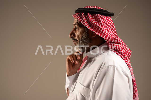 النظر الى شيء ما بإيماءات الجدية، التقدم بالعمر والهيبة والوقار، بورتريه مقرب من الجانب لرجل كبير بالسن عربي خليجي سعودي يرتدي الثوب التقليدي و الشماغ, الاهتمام بالأناقة والمظهر الخارجي، الوقوف باستقامة وثبات، مفهوم الرجولة العربية, خلفية بيج