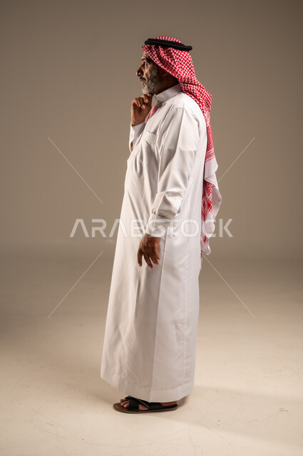 النظر الى شيء ما، التقدم بالعمر والهيبة، بورتريه من الجانب لرجل كبير بالسن عربي خليجي سعودي يرتدي الثوب التقليدي و الشماغ, الاهتمام بالأناقة والمظهر الخارجي، الوقوف باستقامة وثبات، مفهوم الرجولة العربية, الجسم بالطول الكامل،خلفية بيج