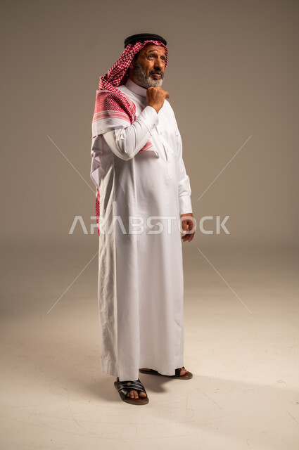 النظر الى شيء ما، التقدم بالعمر والهيبة، بورتريه من الجانب لرجل كبير بالسن عربي خليجي سعودي يرتدي الثوب التقليدي و الشماغ, الاهتمام بالأناقة والمظهر الخارجي، الوقوف باستقامة وثبات، مفهوم الرجولة العربية, الجسم بالطول الكامل،خلفية بيج