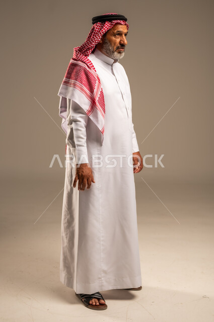 النظر الى شيء ما، التقدم بالعمر والهيبة، بورتريه من الجانب لرجل كبير بالسن عربي خليجي سعودي يرتدي الثوب التقليدي و الشماغ, الاهتمام بالأناقة والمظهر الخارجي، الوقوف باستقامة وثبات، مفهوم الرجولة العربية, الجسم بالطول الكامل،خلفية بيج
