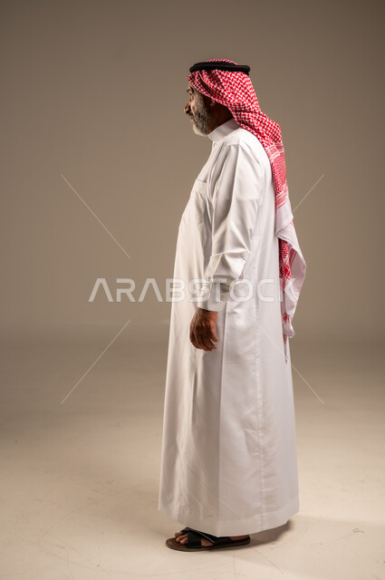 النظر الى شيء ما، التقدم بالعمر والهيبة، بورتريه من الجانب لرجل كبير بالسن عربي خليجي سعودي يرتدي الثوب التقليدي و الشماغ, الاهتمام بالأناقة والمظهر الخارجي، الوقوف باستقامة وثبات، مفهوم الرجولة العربية, الجسم بالطول الكامل،خلفية بيج