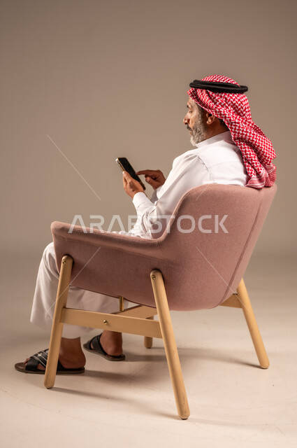 اتصالات ومكالمات هاتفية عبر الهاتف المحمول, الدردشة والتواصل مع الأهل والأصدقاء ، بورتريه من الجانب لرجل كبير بالسن عربي خليجي سعودي يرتدي الثوب التقليدي والشماغ يجلس على كرسي ويستخدم الجوال الهاتفي، خلفية بيج