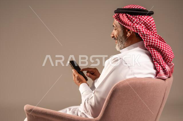 اتصالات ومكالمات هاتفية عبر الهاتف المحمول, الدردشة والتواصل مع الأهل والأصدقاء ، بورتريه من الجانب لرجل كبير بالسن عربي خليجي سعودي يرتدي الثوب التقليدي والشماغ يجلس على كرسي ويستخدم الجوال الهاتفي، خلفية بيج