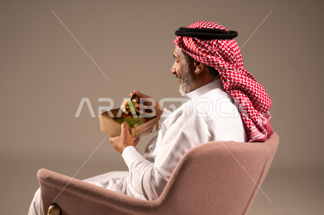 تناول الوجبات الغذائية، بورتريه من الجانب لرجل كبير بالسن خليجي عربي سعودي يجلي على كرسي يحمل طبق من الغذاء، الاستمتاع بالوجبات السريعة، عدم الالتزام بحمية غذائية، خلفية بيج