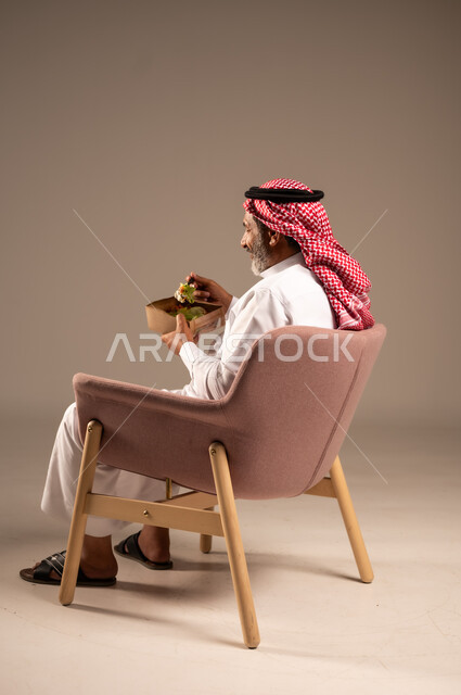 تناول الوجبات الغذائية، بورتريه من الجانب لرجل كبير بالسن خليجي عربي سعودي يجلي على كرسي يحمل طبق من الغذاء، الاستمتاع بالوجبات السريعة، عدم الالتزام بحمية غذائية، خلفية بيج