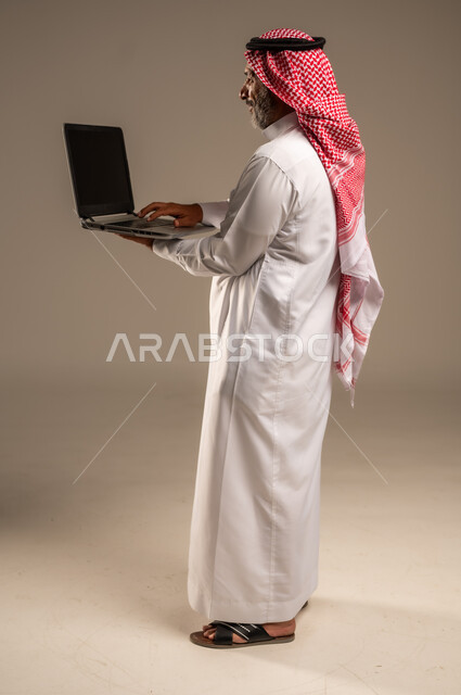 استخدام الحاسوب المحمول لإنجاز المهام، بورتريه لرجل كبير السن عربي سعودي خليجي يرتدي الشماغ والثوب التقليدي يحمل بيده جهاز اللابتوب، تطور التكنولوجيا والتقنيات الحديثة، إجتماع عمل عن بعد، إجراء مكالمات الفيديو, صورة الجسم بالطول الكامل، خلفية بيج
