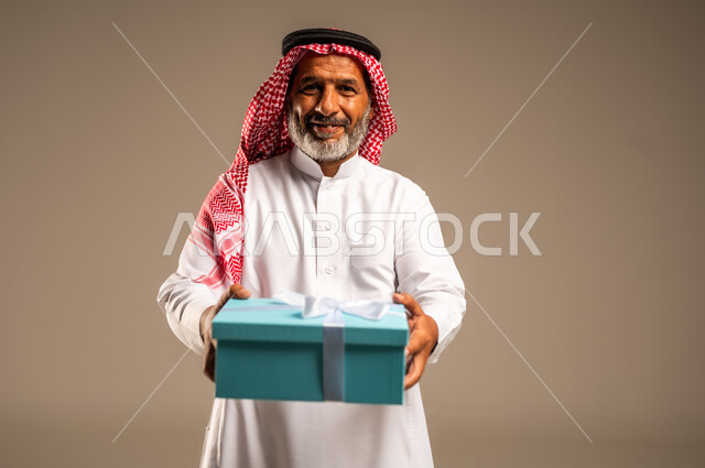 ايماءات وجه تدل على الحماس لمعرفه ما بداخل صندوق الهدايا، بورتريه لرجل كبير بالسن عربي خليجي سعودي  يرتدي الثوب التقليدي والشماغ يمسك بيده هديه كرتونيه، تعابير فرح وسرور واكتشاف المفاجات السعيده، هدايا عيد الاب ومناسبات سعيده، خلفيه بيج