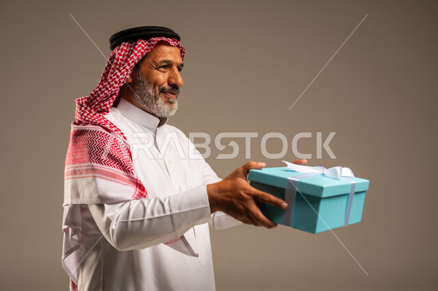 ايماءات وجه تدل على الحماس لمعرفه ما بداخل صندوق الهدايا، بورتريه لرجل كبير بالسن عربي خليجي سعودي  يرتدي الثوب التقليدي والشماغ يمسك بيده هديه كرتونيه، تعابير فرح وسرور واكتشاف المفاجات السعيده، هدايا عيد الاب ومناسبات سعيده، خلفيه بيج
