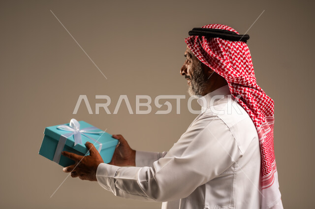 ايماءات وجه تدل على الحماس لمعرفه ما بداخل صندوق الهدايا، بورتريه لرجل كبير بالسن عربي خليجي سعودي  يرتدي الثوب التقليدي والشماغ يمسك بيده هديه كرتونيه، تعابير فرح وسرور واكتشاف المفاجات السعيده، هدايا عيد الاب ومناسبات سعيده، خلفيه بيج