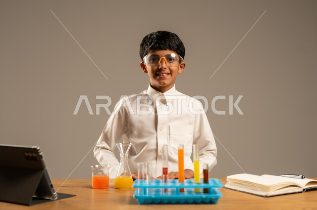 Effective learning methods using technology and technological devices, using experimental educational tools and means, portrait of a Saudi Arabian Gulf student wearing school uniform and safety glasses sitting behind an examination table and sample tubes and chemical analyses, gray background