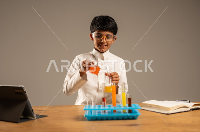 Effective learning methods using technology and technological devices, using experimental educational tools and means, portrait of a Saudi Arabian Gulf student wearing school uniform and safety glasses sitting behind an examination table and sample tubes and chemical analyses, gray background