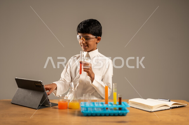 أساليب التعلم الفعال بالاستفادة من التقنيات والأجهزة التكنولوجية، استخدام الأدوات والوسائل التعليمية التجريبية، بورتريه لطالب عربي خليجي سعودي يرتدي الزي المدرسي ونظارات الحماية يجلس خلف طاولة الاختبارات وأنابيب العينات والتحاليل الكيميائية، خلفية رمادية