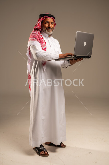 استخدام الحاسوب المحمول لإنجاز المهام، بورتريه لرجل كبير السن عربي سعودي خليجي يرتدي الشماغ والثوب التقليدي يحمل بيده جهاز اللابتوب ينظر الى الكاميرا بإيماءات السرور، تطور التكنولوجيا والتقنيات الحديثة، إجتماع عمل عن بعد، صورة الجسم بالطول الكامل، خلفية بيج