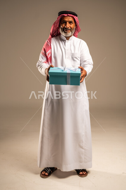 تعابير فرح وسرور واكتشاف المفاجات السعيده، ايماءات وجه تدل على الحماس لمعرفه ما بداخل صندوق الهدايا، بورتريه لرجل كبير بالسن عربي خليجي سعودي  يرتدي الثوب التقليدي والشماغ يمسك بيده هديه كرتونيه، هدايا عيد الاب ومناسبات سعيده، الجسم بالطول الكامل، خلفيه بيج