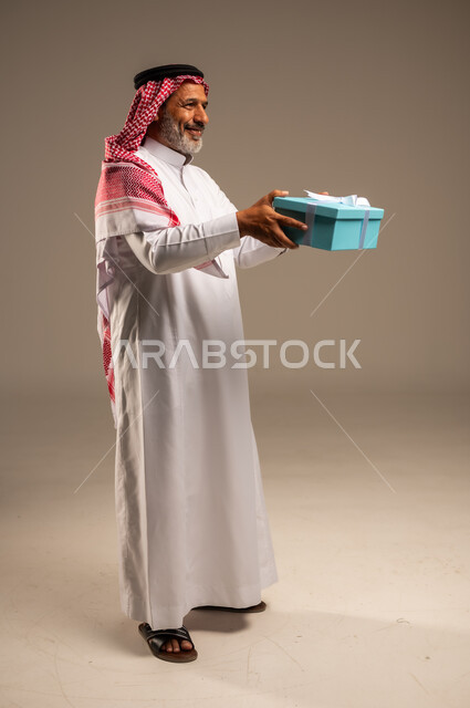 Expressions of joy and happiness and discovering happy surprises, facial gestures indicating excitement to know what is inside the gift box, portrait of an elderly Saudi Arabian Gulf man wearing traditional thobe and shemagh holding a cartoon gift in his hand, Father's Day gifts and happy occasions, full body, beige background