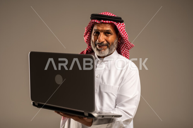 تطور التكنولوجيا والتقنيات الحديثة، استخدام الحاسوب المحمول لإنجاز المهام، بورتريه مقرب لرجل كبير السن عربي سعودي خليجي يرتدي الشماغ والثوب التقليدي يحمل بيده جهاز اللابتوب ينظر الى الكاميرا بإيماءات السرور، إجتماع عمل عن بعد، خلفية بيج