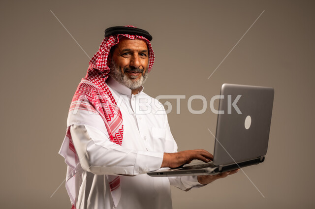 Development of technology and modern techniques, using laptop to accomplish tasks, close-up portrait of elderly Saudi Arabian Gulf man wearing traditional keffiyeh and thobe holding laptop in hand looking at camera with gestures of pleasure, remote business meeting, beige background