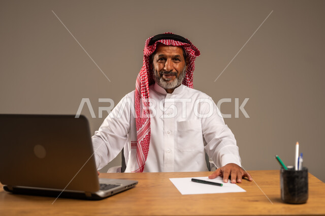 استخدام التقنيات الحديثة والمتطورة في العمل، بورتريه لرجل كبير السن عربي خليجي سعودي يرتدي الثوب التقليدي والشماغ  يجلس خلف طاولة خشبية يعمل على جهاز الحاسوب, خلفية بيج