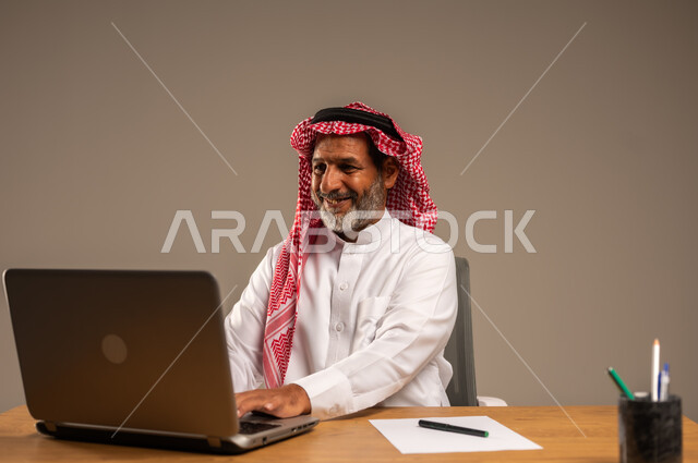استخدام التقنيات الحديثة والمتطورة في العمل، بورتريه لرجل كبير السن عربي خليجي سعودي يرتدي الثوب التقليدي والشماغ  يجلس خلف طاولة خشبية يعمل على جهاز الحاسوب, خلفية بيج