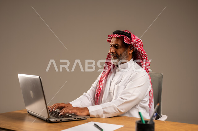 استخدام التقنيات الحديثة والمتطورة في العمل، بورتريه لرجل كبير السن عربي خليجي سعودي يرتدي الثوب التقليدي والشماغ  يجلس خلف طاولة خشبية يعمل على جهاز الحاسوب, خلفية بيج