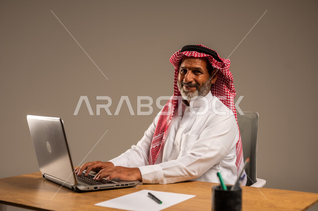 استخدام التقنيات الحديثة والمتطورة في العمل، بورتريه لرجل كبير السن عربي خليجي سعودي يرتدي الثوب التقليدي والشماغ  يجلس خلف طاولة خشبية يعمل على جهاز الحاسوب, خلفية بيج