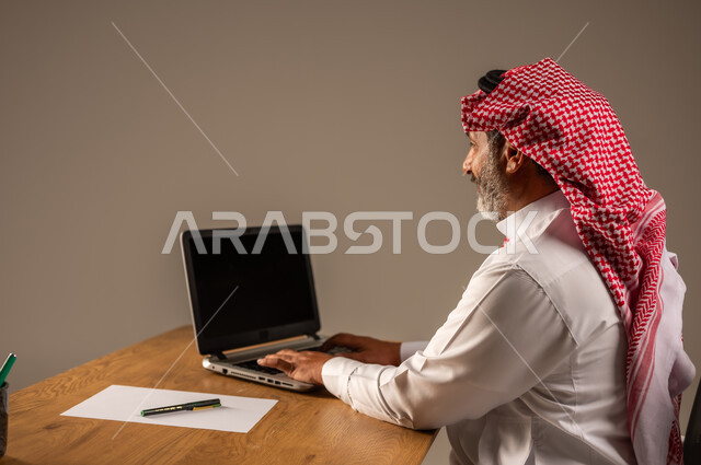 Using modern and advanced technologies in work, portrait of an elderly Saudi Arabian Gulf man wearing traditional thobe and shemagh sitting behind a wooden table working on a computer, beige background