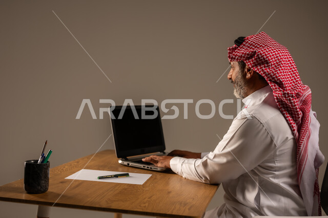 استخدام التقنيات الحديثة والمتطورة في العمل، بورتريه لرجل كبير السن عربي خليجي سعودي يرتدي الثوب التقليدي والشماغ  يجلس خلف طاولة خشبية يعمل على جهاز الحاسوب, خلفية بيج
