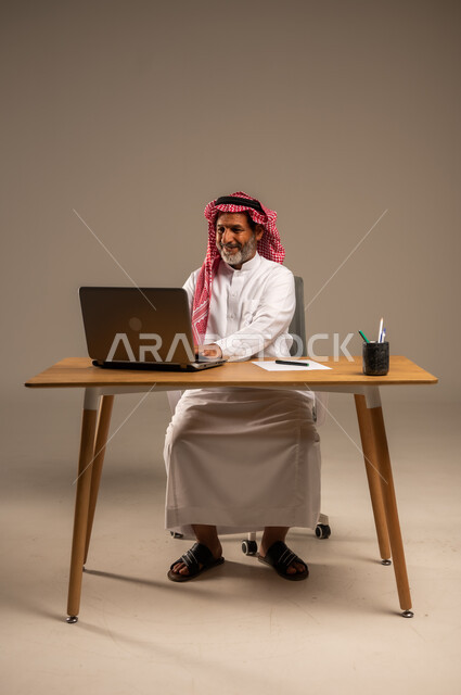 مهن ووظائف مكتبيه سعودية، استخدام التقنيات الحديثة والمتطورة في العمل، بورتريه لرجل كبير السن عربي خليجي سعودي يرتدي الثوب التقليدي والشماغ  يجلس خلف طاولة خشبية يعمل على جهاز الحاسوب, الجسم بالطول الكامل، خلفية بيج