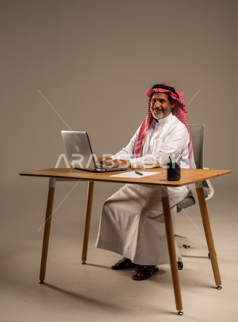 مهن ووظائف مكتبيه سعودية، استخدام التقنيات الحديثة والمتطورة في العمل، بورتريه لرجل كبير السن عربي خليجي سعودي يرتدي الثوب التقليدي والشماغ  يجلس خلف طاولة خشبية يعمل على جهاز الحاسوب, الجسم بالطول الكامل، خلفية بيج