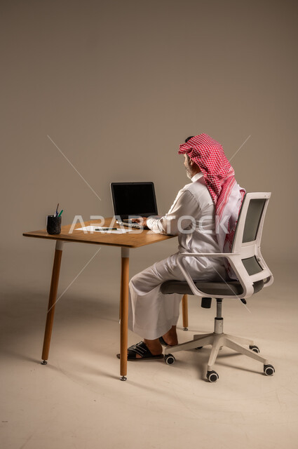 Saudi office jobs and professions, using modern and advanced technologies in work, portrait of an elderly Saudi Arabian Gulf man wearing traditional thobe and shemagh sitting behind a wooden table working on a computer, full body, beige background